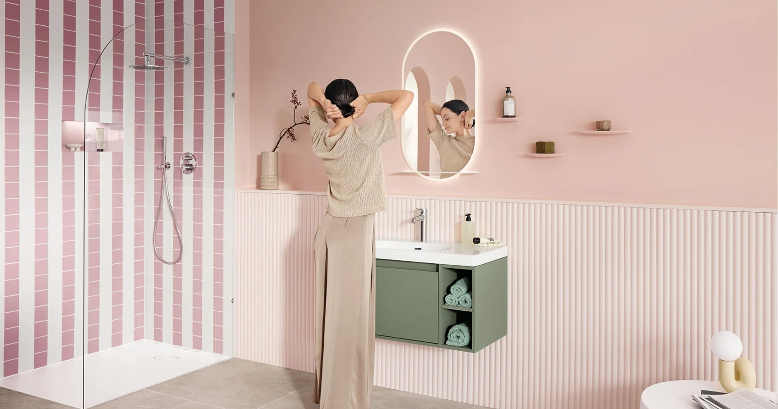 A woman stretches in front of the mirror at the wash area in a stylishly furnished pastel pink bathroom. The wash area consists of a white washbasin with a delicate green base cabinet and the faucet in chrome from the Skyla collection. The illuminated, oval More To See Lite+ mirror has a practical shelf in white.