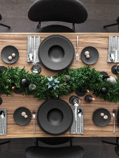 A top view of a Christmas dining table setting with black plates from the Villeroy & Boch Manufacture range together with Piemont cutlery. It is also decorated with a centrepiece of green foliage and candles on a wooden surface.