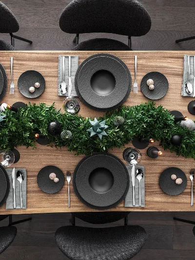 A top view of a Christmas dining table setting with black plates from the Villeroy & Boch Manufacture range together with Piemont cutlery. It is also decorated with a centrepiece of green foliage and candles on a wooden surface.