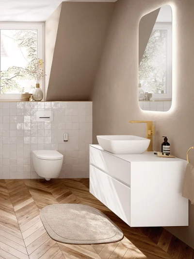 Nowoczesna beżowa łazienka z toaletą z funkcją prysznica i nablatową umywalką Nowoczesna beżowa łazienka z białą toaletą ViClean I200, nablatową umywalką Artis umieszczoną na szafce Collaro, złotą baterią i podświetlanym lustrem nad drewnianą podłogą w jodełkę.
