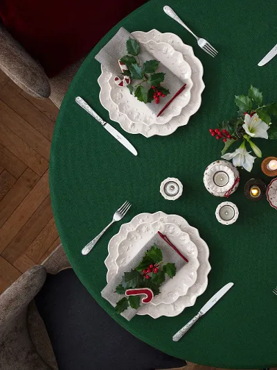 Runder, grün gedeckter Tisch, dekoriert mit dem Geschirr Toy’s Delight Royal Classic von Villeroy & Boch Ein runder Tisch für vier Personen mit weißen Tellern von Villeroy & Boch Toy’s Delight Royal Classic, einer grünen Tischdecke und weihnachtlichen Dekorationen, darunter Stechpalmenblätter, Beeren und Zuckerstangen, von oben betrachtet.
