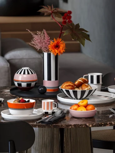 Memphis Breakfast Table Setting Modern breakfast table styled with Memphis porcelain featuring bold black-and-white stripes, pink and orange accents, pastries, fruit, and a striped vase with vibrant floral arrangement.