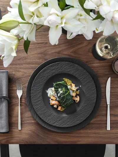 Manufacture by Villeroy & Boch for special occasions Elegant table setting with black Manufacture plates, cutlery, figs, a dish with vegetables and prawns, grey napkins, white flowers, a wine glass and sliced bread.