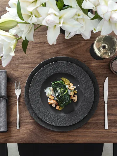 Manufacture by Villeroy & Boch for special occasions Elegant table setting with black Manufacture plates, cutlery, figs, a dish with vegetables and prawns, grey napkins, white flowers, a wine glass and sliced bread.