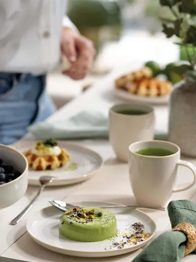 Person preparing a breakfast table with various dishes on Crafted Cotton breakfast plates and with cups of tea.