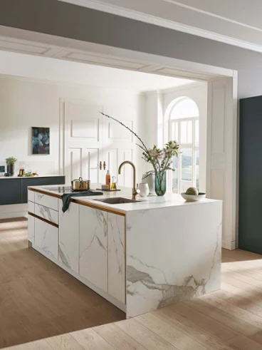 Golden Umbrella kitchen tap with marble kitchen island A modern kitchen with marble island, wooden floor, dark-coloured cabinets, built-in oven and large window. The furnishings include a vase with flowers, kitchen utensils and a houseplant on the windowsill.