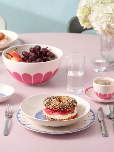 A round pink table with plates full of food, including a filled bagel, a bowl of fruit, cups of coffee and cutlery. A round pink table with plates full of food, including a filled bagel, a bowl of fruit, cups of coffee and cutlery. In the background is a vase of white flowers.