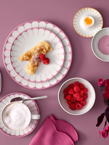Vue de dessus à plat d'assiettes en porcelaine rose Fleur de Villeroy & Boch avec une crêpe, d'une tasse de cappuccino, d'un beurrier et de petits bols remplis de framboises fraîches et d'un œuf à la coque, sur un fond rose poudré à côté d'un feuillage magenta.