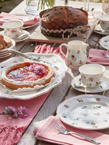 Petite Fleur dinnerware on a coffee table The Petite Fleur collection from Villeroy & Boch is elegantly arranged on a wooden table and includes plates with a floral pattern, a cake, a tart, cups, and pink napkins for tea.