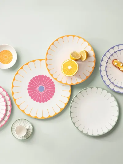 Various colourful, patterned plates, bowls and a cup are arranged on a bright surface. Various colourful, patterned plates, bowls and a cup are arranged on a light-coloured surface, each with a different design. Pastries are placed on some of the plates.
