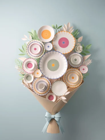 A floral arrangement of kitchenware with various plates and bowls decorated with fruit motifs, wrapped in brown paper and tied with a light blue ribbon, against a light blue background.