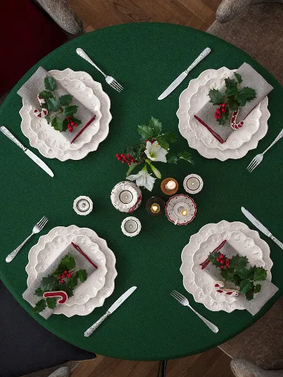 Runder, grün gedeckter Tisch, dekoriert mit dem Geschirr Toy’s Delight Royal Classic von Villeroy & Boch Ein runder Tisch für vier Personen mit weißen Tellern von Villeroy & Boch Toy’s Delight Royal Classic, einer grünen Tischdecke und weihnachtlichen Dekorationen, darunter Stechpalmenblätter, Beeren und Zuckerstangen, von oben betrachtet.