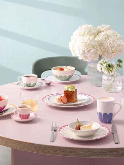 A round table with plates, cups and bowls full of food, decorated with pink patterns. In the centre is a vase of white flowers and two unlit candles. Chairs surround the table.	