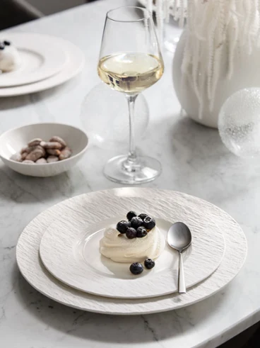 A dessert on a Manufacture plate A dessert on a Manufacture plate garnished with berries, a spoon, a wine glass, and a bowl of nuts on a white tablecloth.