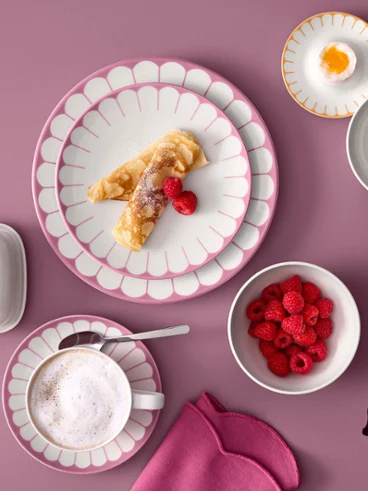 Vue d'en haut d'une élégante table de petit-déjeuner présentant la collection Fleur de Villeroy & Boch aux tons rose pastel, vert menthe et orange. Comprend des assiettes avec des crêpes et des framboises, une tasse de cappuccino, un beurrier, un coquetier et un bol de framboises fraîches sur un fond rose poudré.