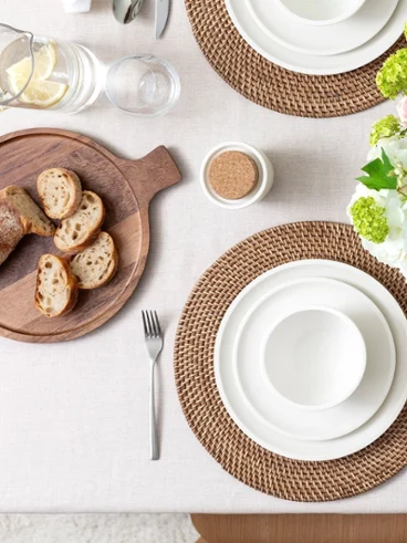 A table set with white dinnerware on rattan placemats with a wooden serving tray with bread on it