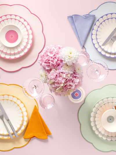 Vier pastellfarbene Gedecke der Fleur Kollektion mit geschwungenen Tellern, Servietten und Besteck umgeben einen Strauß rosa Blumen.
