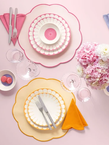 Four pastel-coloured place settings from the Fleur collection with curved plates, napkins and cutlery surround a bouquet of pink flowers.