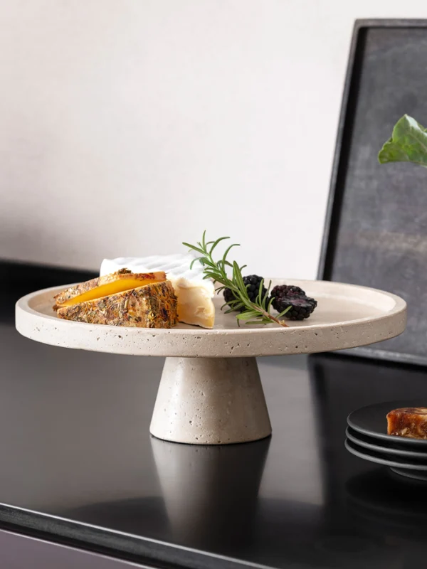 Plat de service parfait en travertin pour antipasti ou fromage. Un assortiment de fromages sur un plat de service en travertin avec pied, et des fruits foncés, à côté d'une vaisselle noire Manufacture de Villeroy & Boch sur un plan de travail foncé.