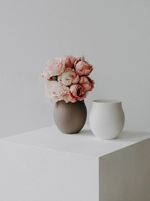 Een bruine vaas met roze bloemen en een witte vaas van keramiek staan op een witte kubus tegen een effen lichtkleurige achtergrond.