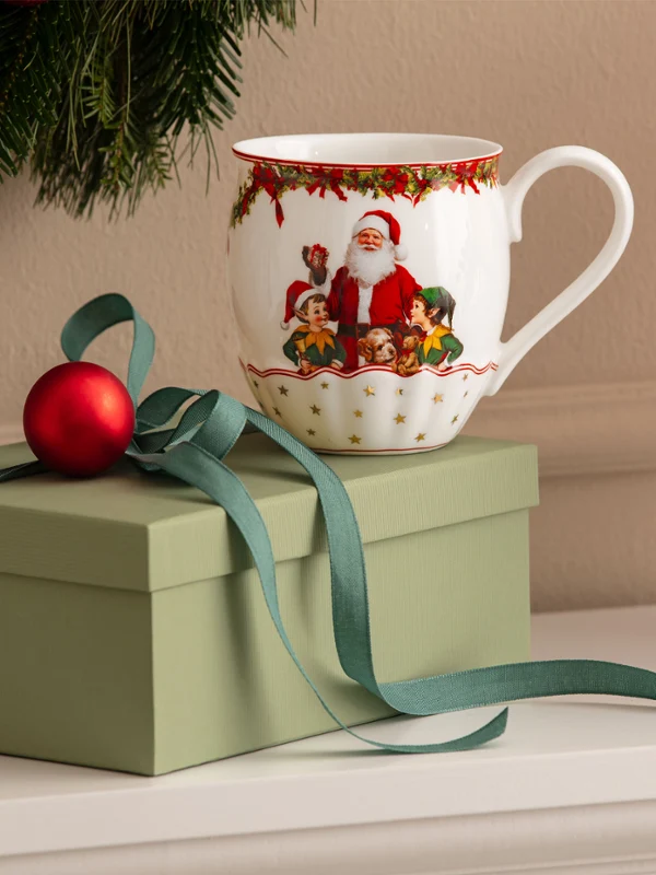 A green gift box with a bow, a red Christmas ornament, and a mug with a Santa Claus motif from Villeroy & Boch in front of a white background, with Christmas decorations visible in the background.