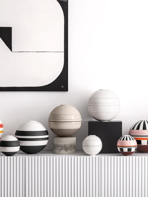A series of patterned spherical tableware sets on a white shelf A series of patterned spherical tableware sets on a white shelf, underneath a geometric black-and-white painting featuring a vase with orange flowers.