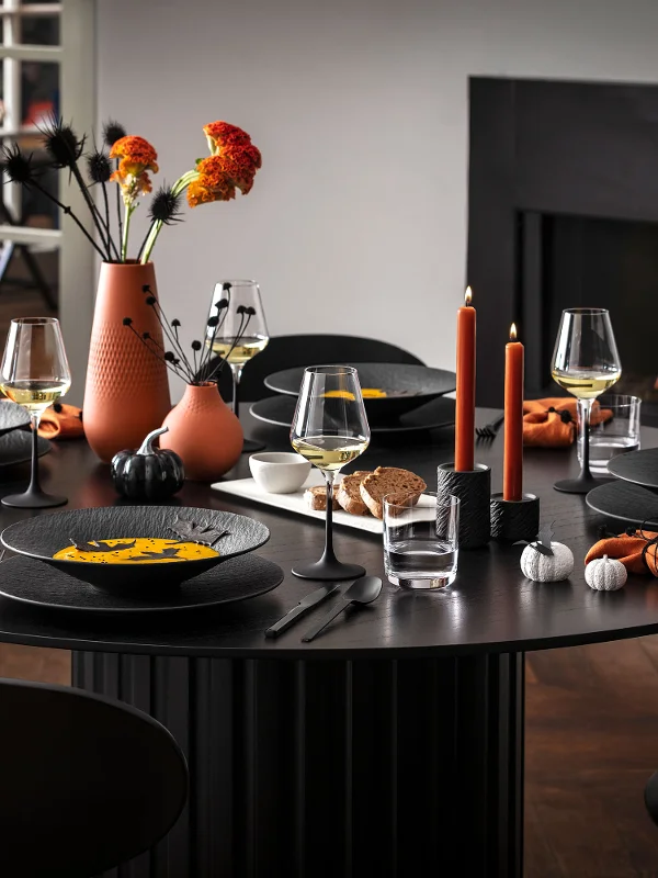 Un moderno tavolo da pranzo per cinque persone con stoviglie nere Manufacture, tovaglioli arancioni e decorazioni autunnali, tra cui vasi di fiori e candele accese, accanto a un camino acceso.