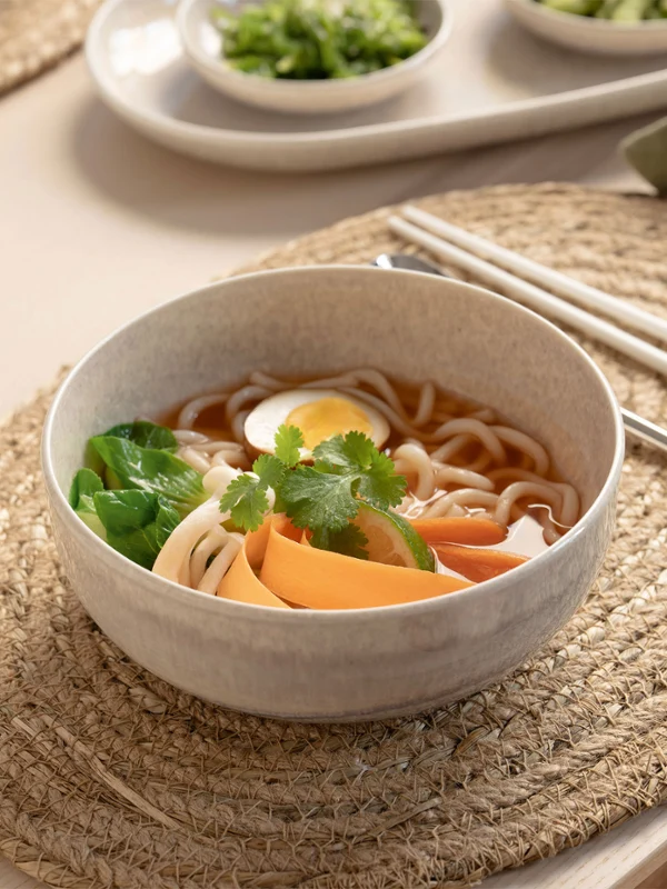 A bowl from the Perlemor collection with a noodle soup A bowl from the Perlemor collection with noodle soup with egg, vegetables and herbs on a woven placemat with chopsticks.