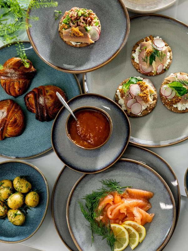Eine Auswahl an Gourmetgerichten, darunter Räucherlachs, belegte Brötchen und Gebäck, serviert auf stilvollen Lave-Tellern von Villeroy & Boch auf einem weißen Tisch.