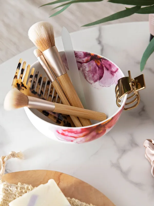 Various make-up brushes in a Rose Garden cereal bowl with a floral decor arranged on a marble tabletop beside decorative objects.