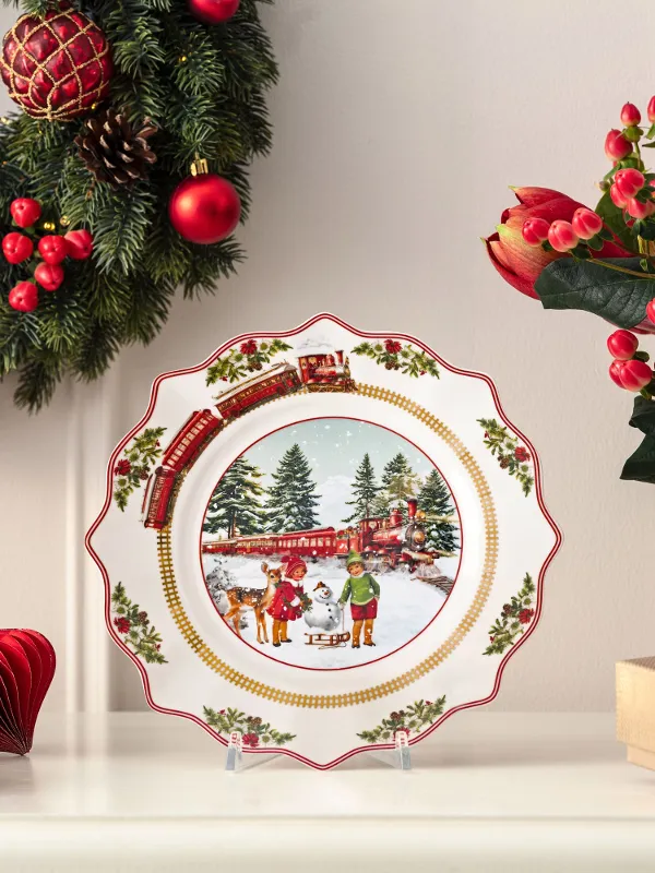 A decorative Christmas plate featuring a winter scene is displayed on a mantelpiece adorned with Christmas garlands, flowers, and wrapped presents.