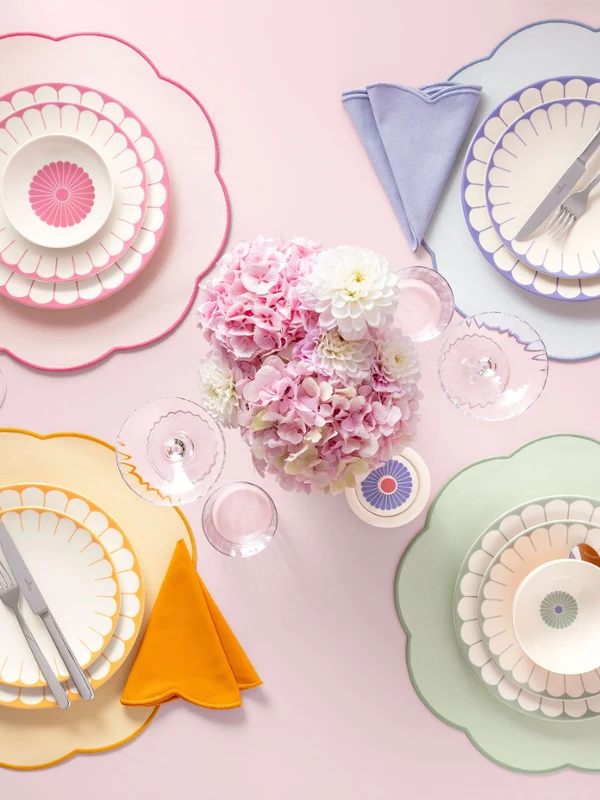 Colorful table setting with colorful Fleur plates, bowls, and placemats Four placemats with colorful plates and bowls from the Fleur collection, colorful napkins, and cutlery are arranged around a pink floral decoration.