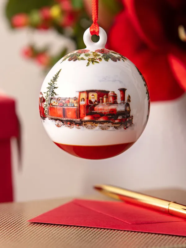 Close-up of a Christmas ornament with a train motif, with wrapped gifts, a red envelope, and a golden pen in the background.