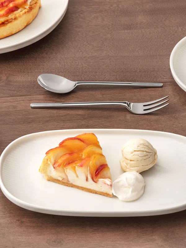 Ein Stück Obsttorte mit zwei Kugeln Eis auf dem Afina Teller, daneben eine Tasse Tee und Besteck ebenfalls aus der Afina Kollektion auf einem Holztisch.