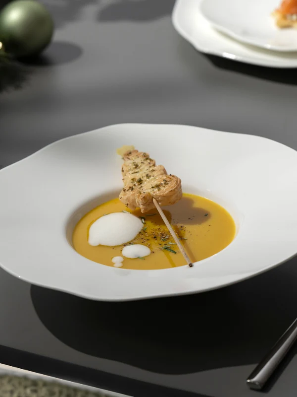 Un bol Pura blanco con sopa, una rebanada de pan y espuma en una mesa puesta con cubiertos y una servilleta.