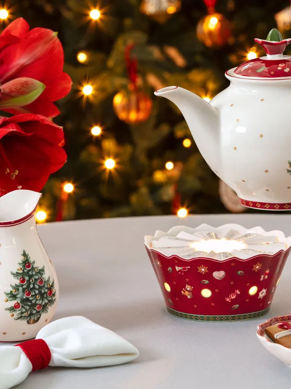 Una tavola festiva con una teiera, uno scaldavivande, una tazza, ceramiche natalizie, biscotti e un albero decorato sullo sfondo.