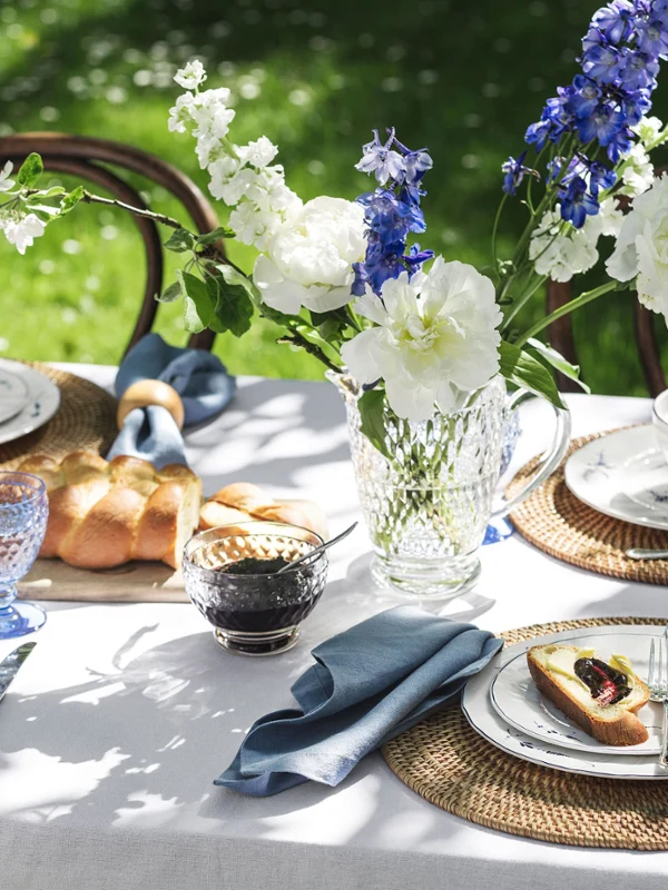 Outdoor table setting with flowers, blue glasses, bread and Old Luxembourg plates with food on woven placemats in a garden.