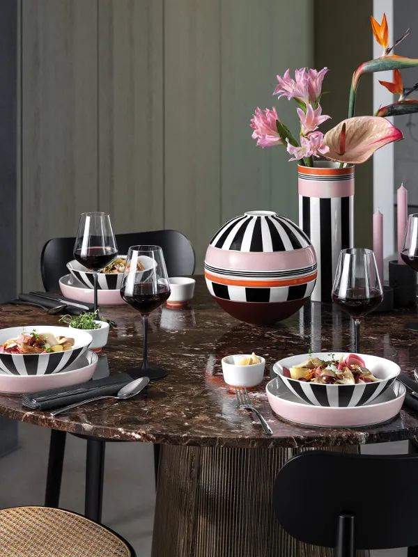 Un tavolo rotondo in marmo apparecchiato per quattro, con stoviglie Memphis a righe, bicchieri da vino e un vaso di fiori rosa.