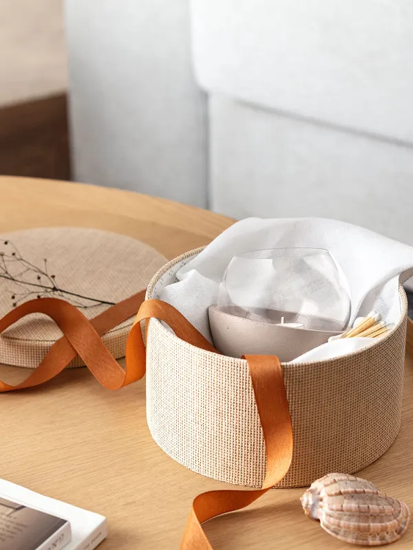 A round woven basket with an orange bow containing a NewMoon Home hurricane lamp on a wooden table next to a black Collier vase.