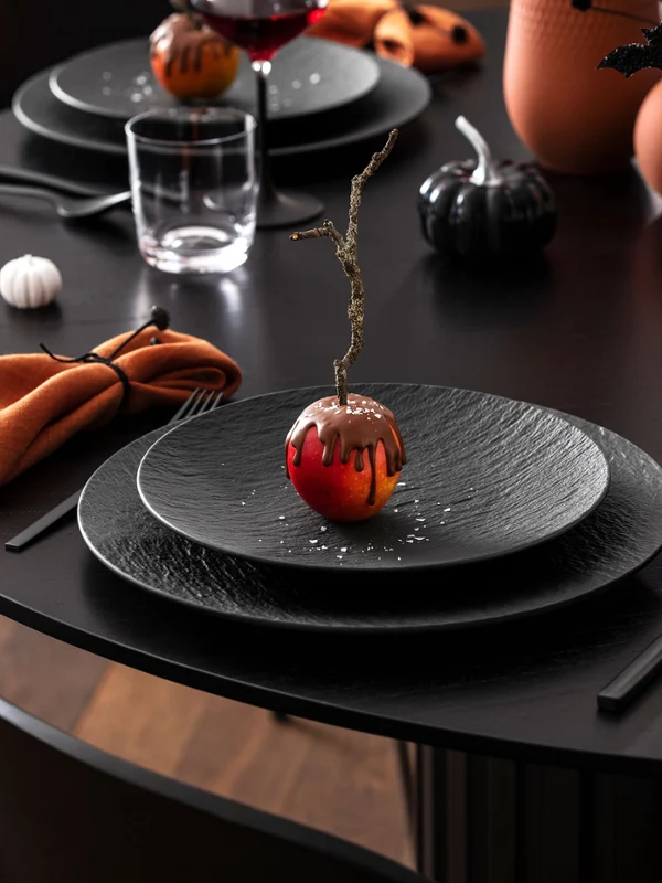 Un tavolo da pranzo nero è apparecchiato con piatti neri Manufacture, tovaglioli arancioni e bevande e decorazioni a tema Halloween, tra cui una mela ricoperta di cioccolato.