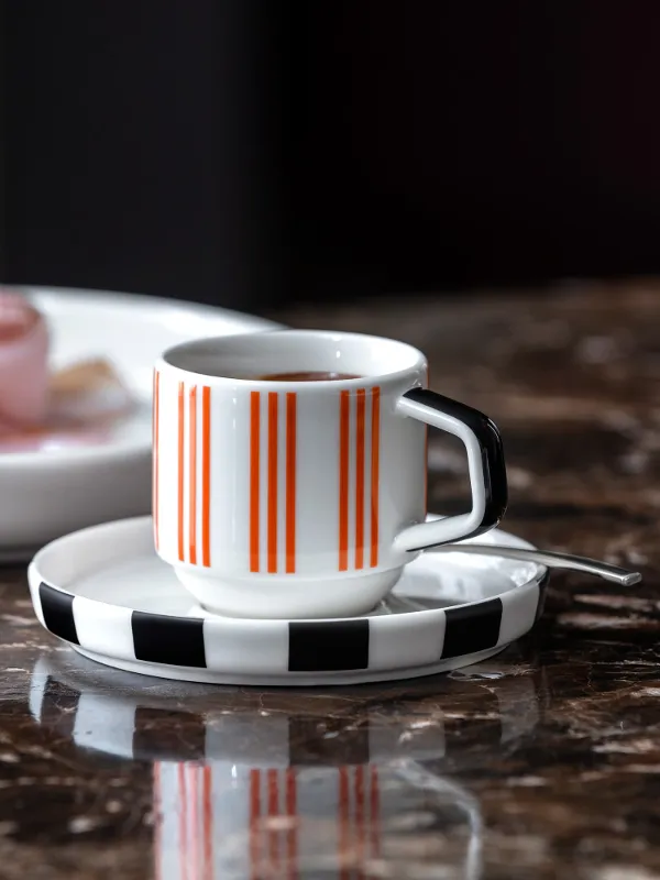 Una tazza da espresso Memphis a righe su un piattino si trova su un tavolo di marmo, accanto a un piatto Memphis.

