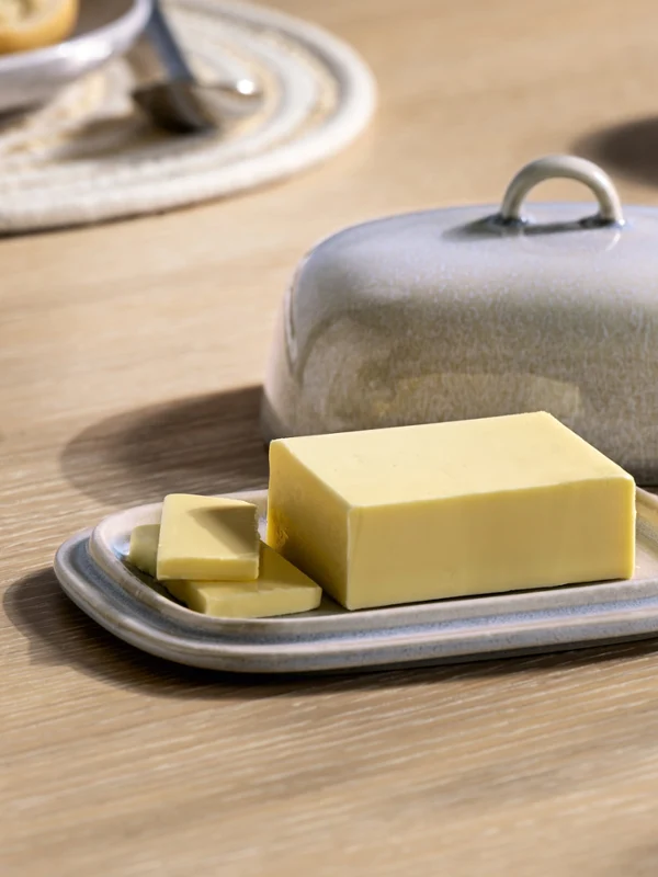 A rectangular block of butter with two slices lies on a ceramic Perlemor butter dish with a matching lid on a wooden table.