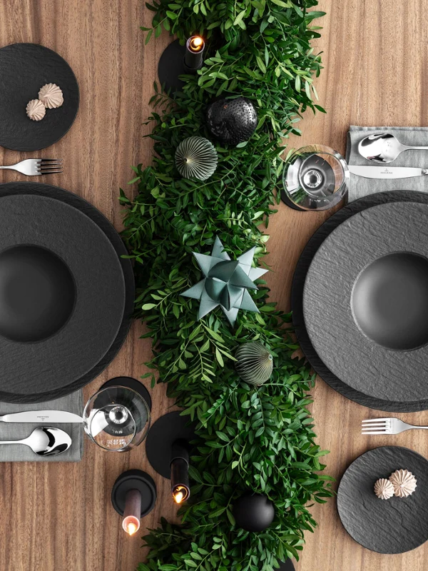 Un tavolo da pranzo splendidamente apparecchiato con piatti neri Manufacture, tovaglioli grigi e posate d'argento. Una decorazione da tavola con fogliame verde lungo il tavolo, accentuata da candele e oggetti decorativi.