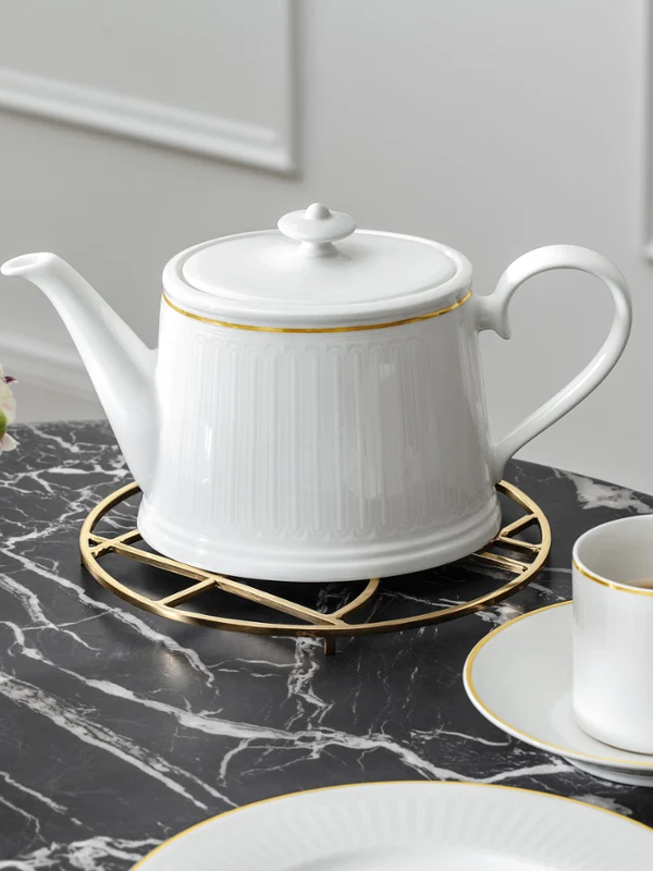 A white coffee pot with a gold rim on a stand on a marble table. Next to it is a matching teacup and saucer with a gold spoon and a black vase with a bouquet of flowers.