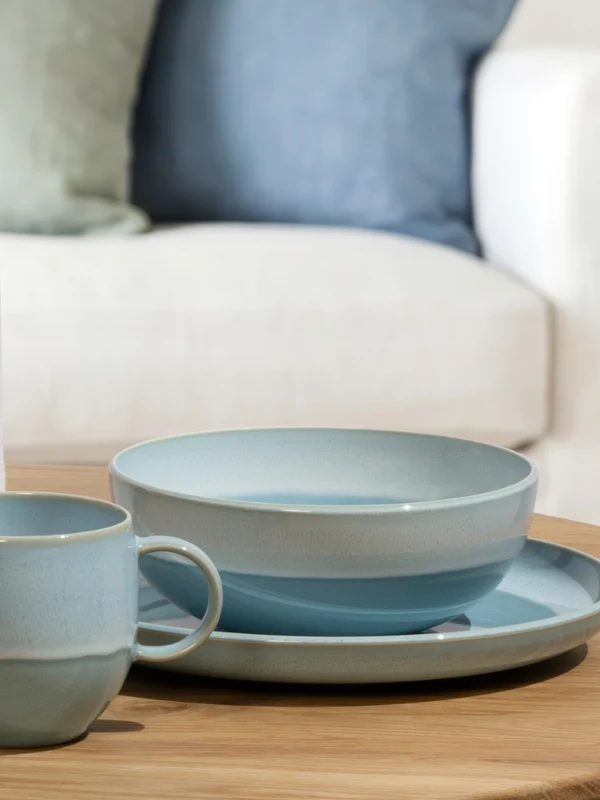 Vajilla azul claro Perlemor y una taza a juego se encuentran sobre una mesa de madera al lado de un regalo envuelto con una rodaja de naranja seca y una cinta.