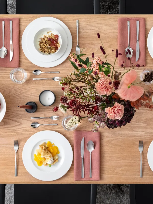 A rectangular wooden table is set for four people. There are plates with food, pink napkins, cutlery, candles, and a floral arrangement on the table. Grapes and spices are also present.