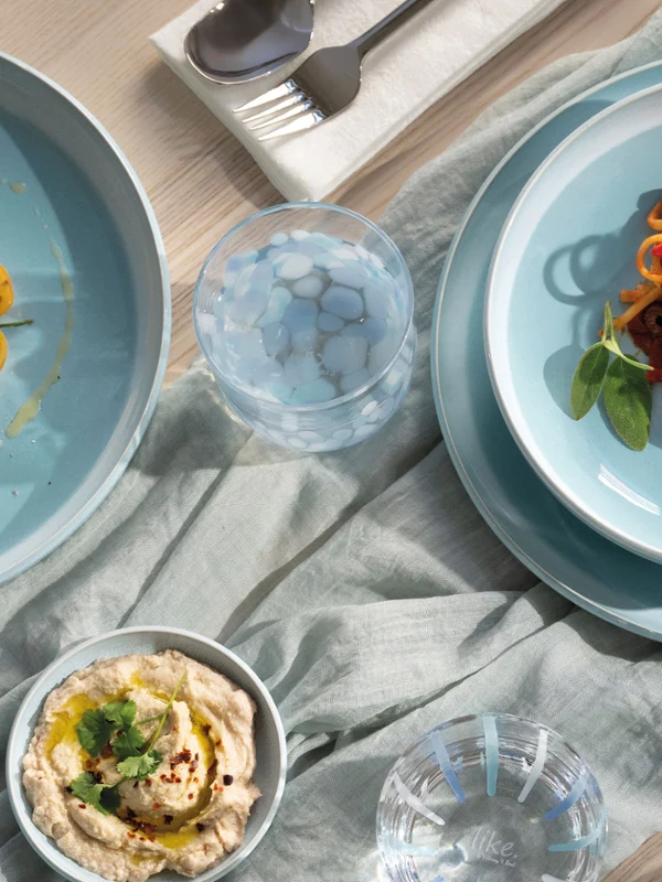 Ein gedeckter Tisch mit Nudeln, Salat, Hummus, Oliven und Utensilien auf blauen Tellern der Perlemor Aqua Kollektion  und einer hellen Tischdecke.
