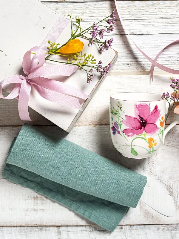 A hand reaches for a Villeroy & Boch Mariefleur mug next to a gift box with a pink ribbon and flowers, placed on a wooden surface with a teal cloth.
