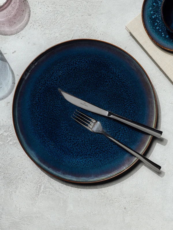 Een mooi gedekte tafel met een donkerblauw Crafted bord, bestek en een boek op een betonnen oppervlak.