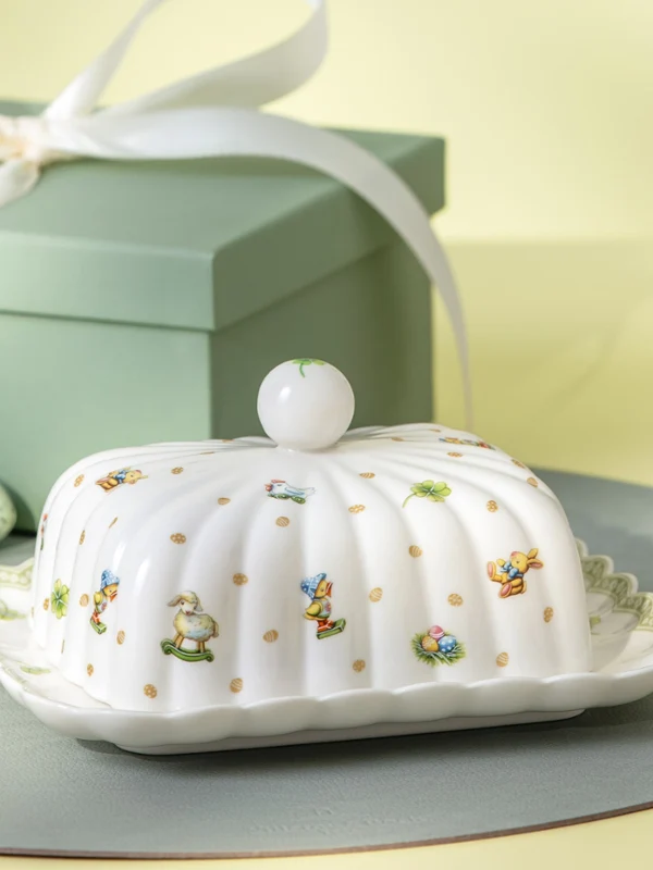 A decorative butter dish by Easter Delight with flowers and Easter patterns stands next to a green gift box and pastel-colored speckled eggs on a table.